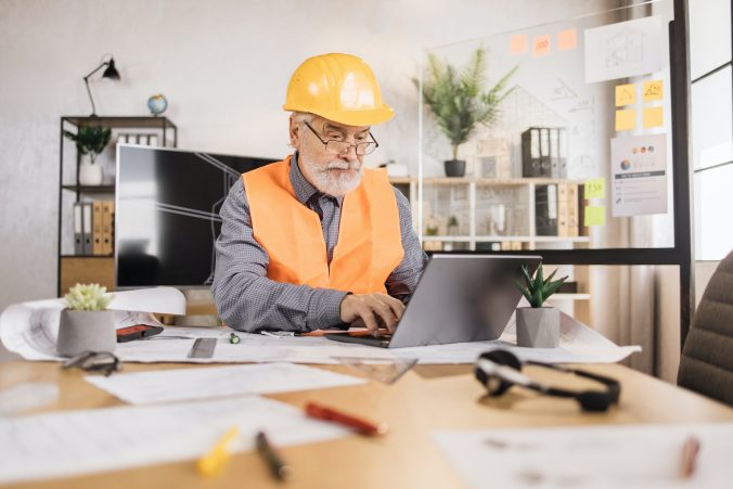 Engineer senior man in helmet and reflective vest sitting at table with lots of blueprints